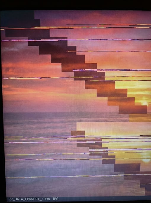 Shattered horizontal displacement bands tear through a corrupted sunset photograph — the original warm oranges and pinks visible but broken into jagged staircase patterns