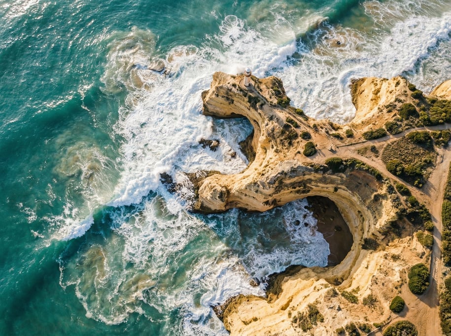Top-down aerial view of a dramatic coastal cliff edge where turquoise ocean waves crash against gold