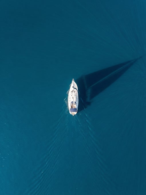 Top-down view of a single white sailboat on deep cerulean blue open water, casting a long shadow