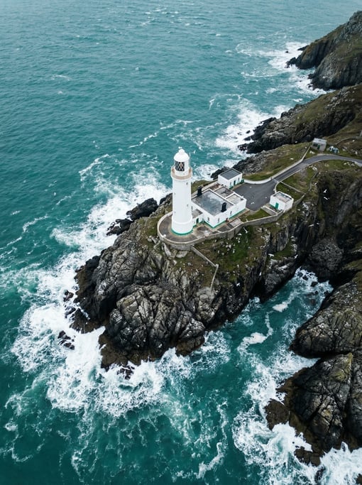 Overhead aerial of a lighthouse on a rocky promontory, white tower against dark grey cliffs