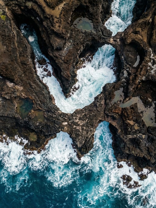 Top-down view of a rocky coastline with waves surging through narrow sea channels carved in dark sto