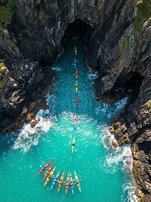 Overhead view of sea kayakers as tiny colorful dots paddling through a narrow sea cave entrance