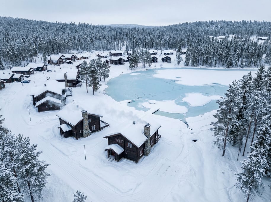 Aerial view of a snowy Nordic village with dark wooden chalets, wisps of chimney smoke