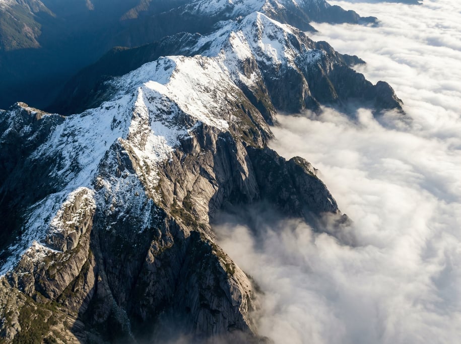 Aerial shot of a mountain ridge from above with snow on the peaks, exposed dark granite rock faces