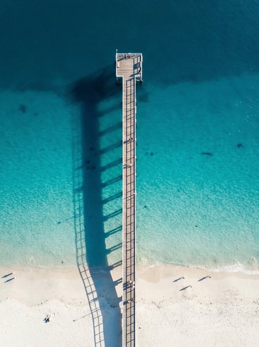 Overhead shot of a wooden pier extending straight out into deep blue ocean from a white sand beach