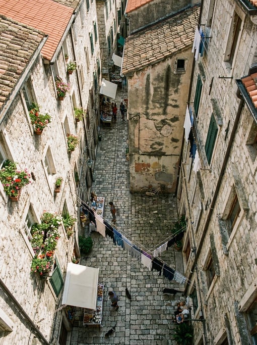 Top-down aerial of a narrow cobblestone alley between tall buildings with flowering window boxes