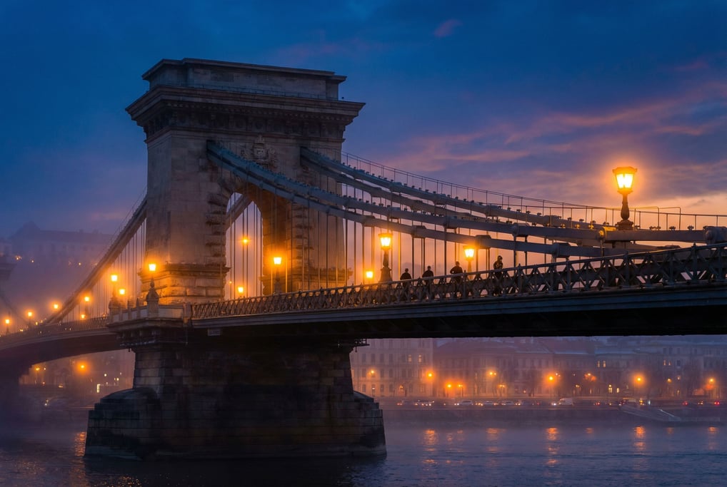 Suspension bridge spanning a wide river at dusk, warm streetlights creating halos in fog