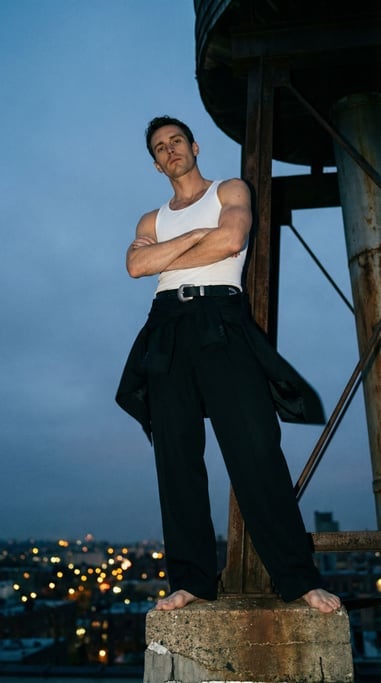 Low-angle editorial shot of a man in a white tank top and black tailored wide-leg trousers with heav