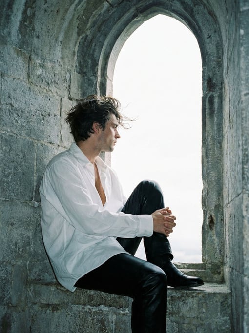 Editorial fashion photograph of a man in a loose white poet shirt unbuttoned to mid-chest and black