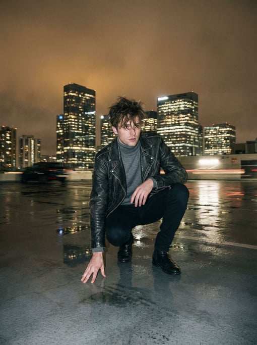 Editorial fashion photograph of a man in a black leather motorcycle jacket over a fine-gauge grey me