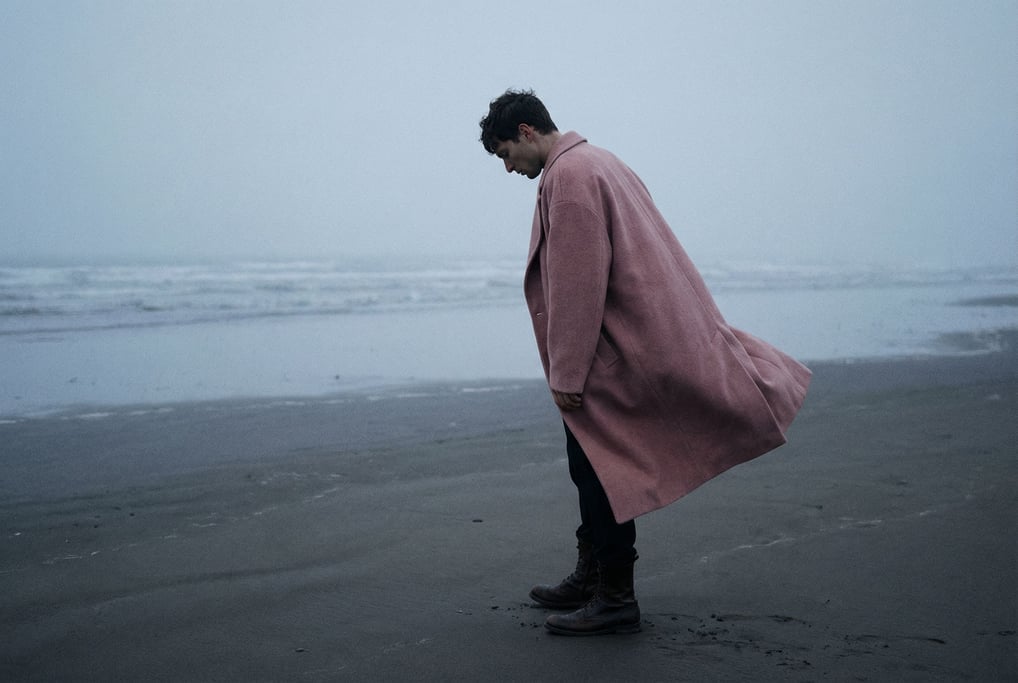 Editorial fashion photograph of a man in a dusty rose oversized wool coat and black boots standing a