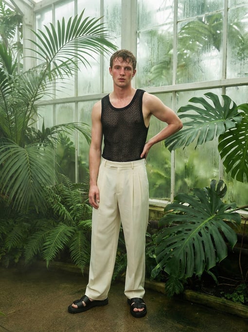 Editorial fashion photograph of a man in a black mesh tank top and wide cream pleated trousers with