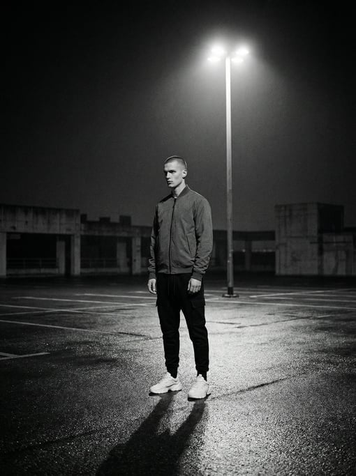 Editorial fashion photograph of a man in a stone-grey technical fabric bomber jacket and black taper