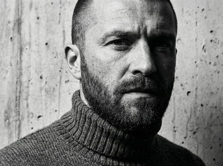 Editorial portrait of a man with close-cropped hair and a full beard in a charcoal ribbed knit turtl