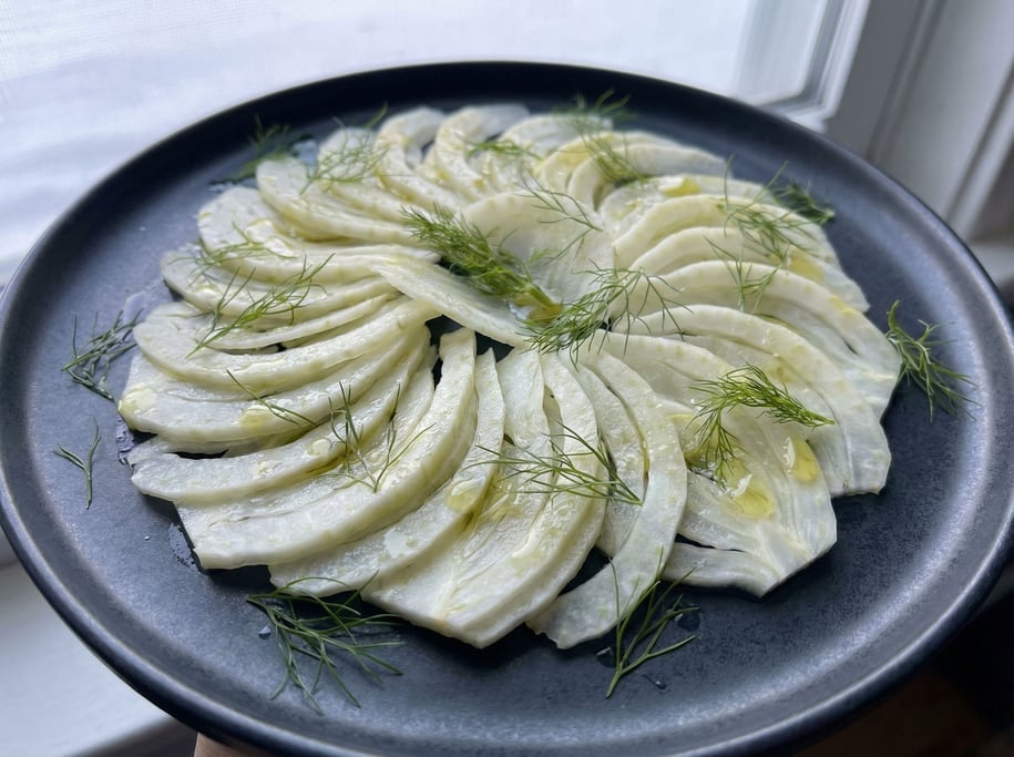 Fennel bulb sliced thin on a mandoline, the translucent white slices fanning across a dark plate