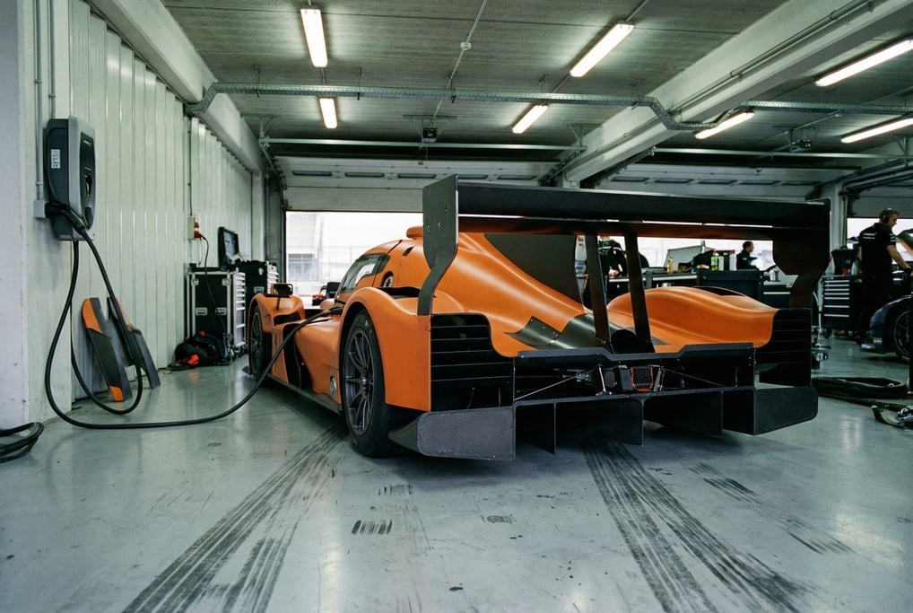 A single-seat racing pod in matte orange with black carbon-fiber aero elements sits in a pit garage