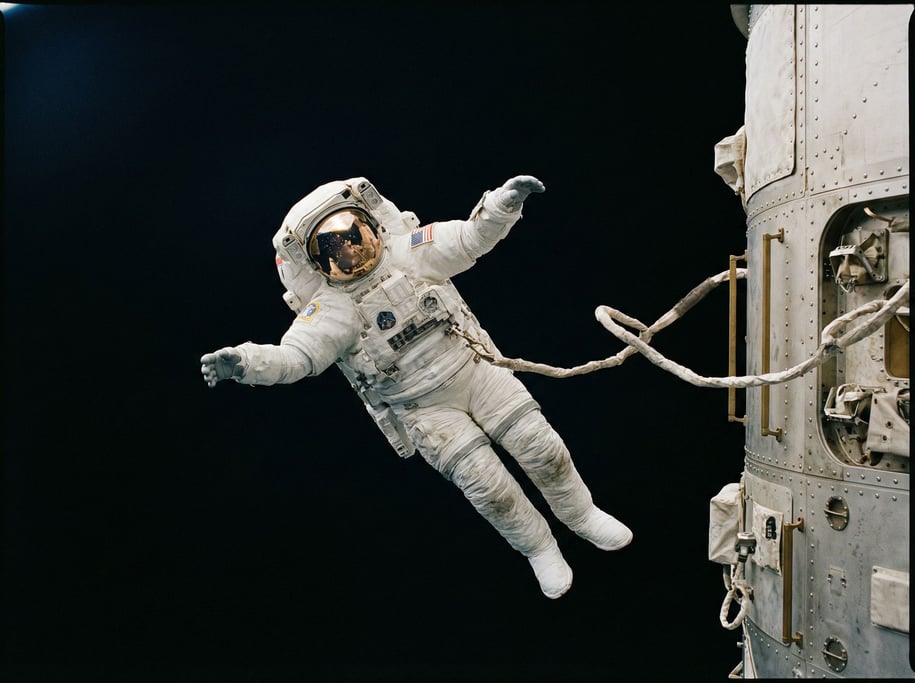 An EVA astronaut floats against the black of space, tethered to a hull visible at the frame's edge