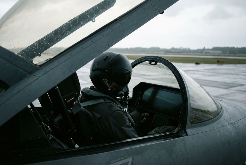 A pilot in a dark flight suit sits in a cockpit seat, seen from the side through an open canopy