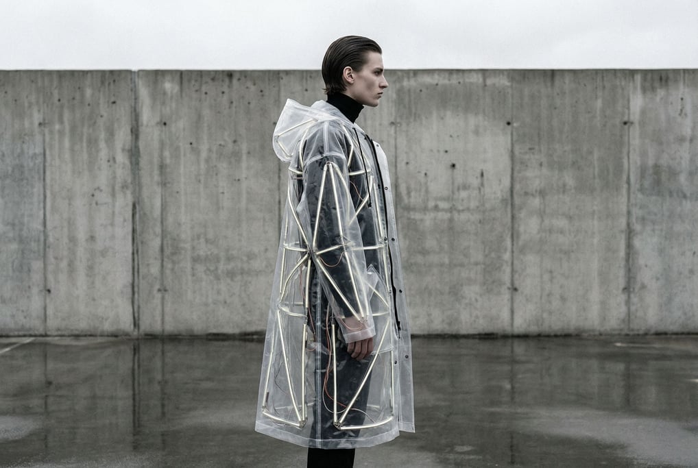 A model stands in profile against a concrete wall wearing an oversized translucent raincoat with geo