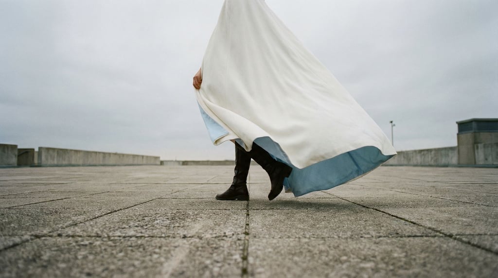 A figure in a billowing white cape walks alone across a concrete plaza in wind