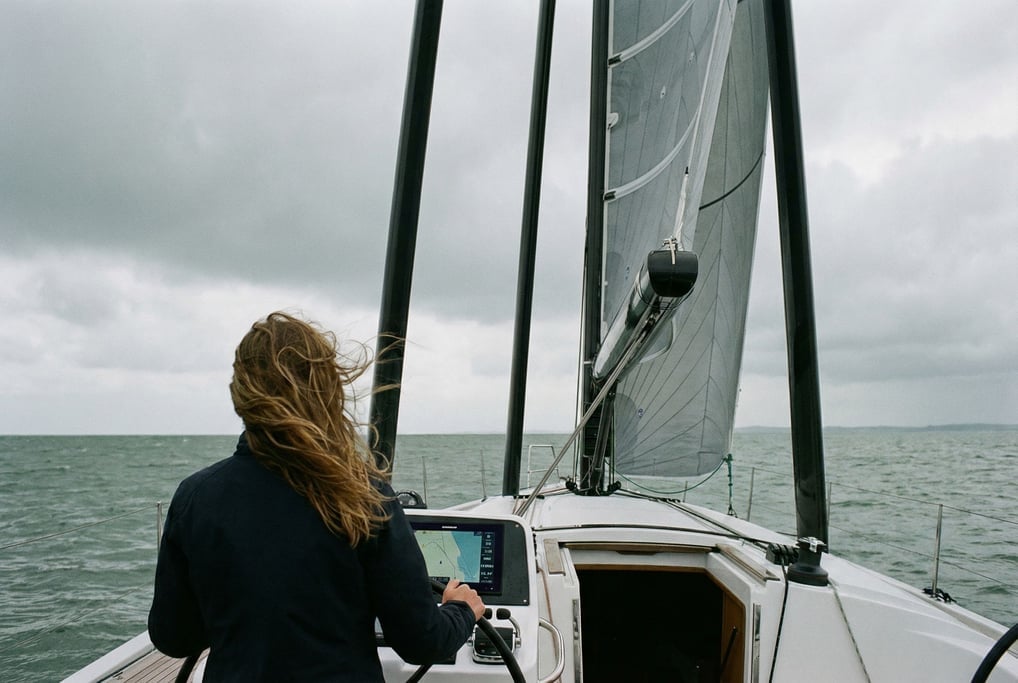 A woman photographed from behind stands at the helm of a sailing yacht with an automated rigging sys