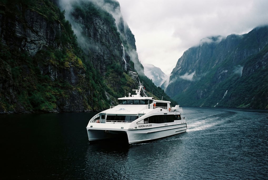 A catamaran ferry in glossy white with an enclosed passenger cabin cruises through a fjord