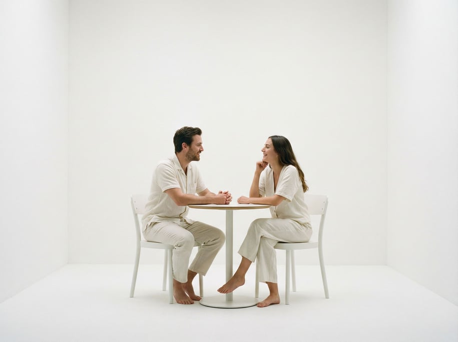 Two people — a man and a woman — sit across from each other at a small round table in a bare white r