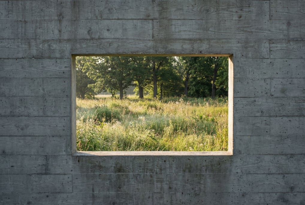 A concrete wall with a precise rectangular opening framing a view of wild meadow and trees beyond