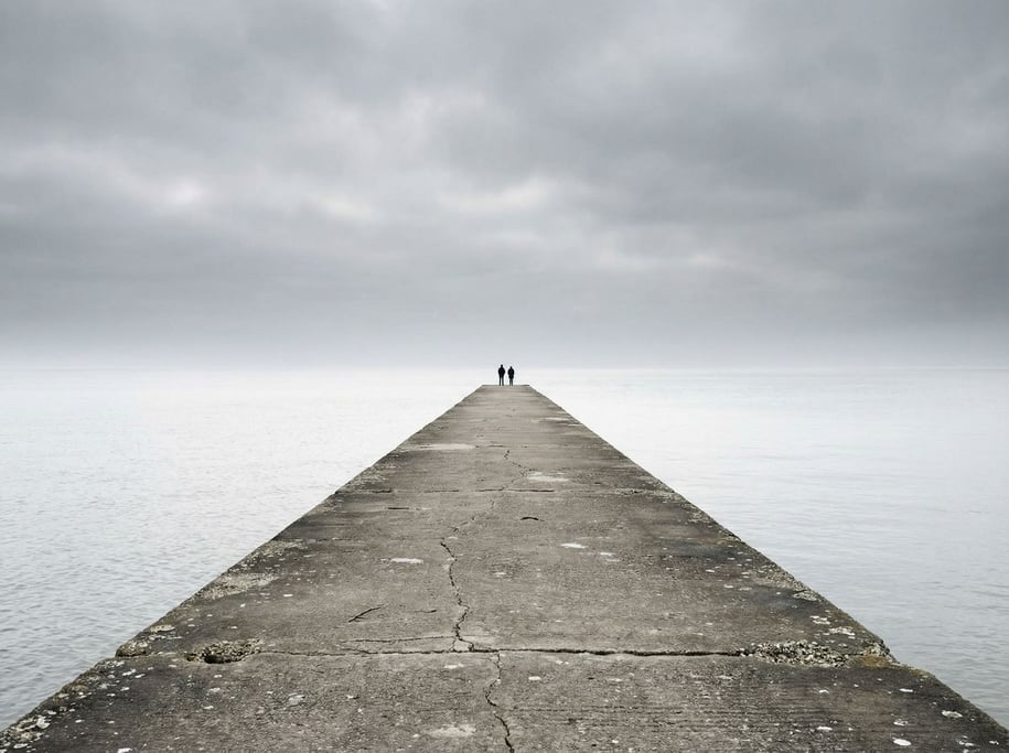 Two small figures at the end of a vast concrete pier extending into calm ocean