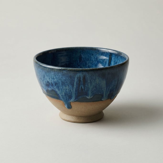 Hand-thrown ceramic bowl with ocean blue drip glaze pooling over a raw natural clay body and foot ri