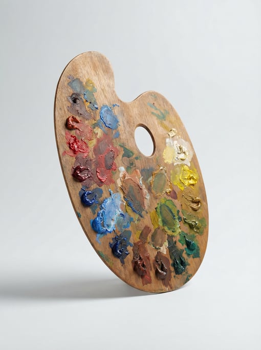Wooden kidney-shaped artist palette with dried remnant dabs of vivid oil paint colors and a thumb ho