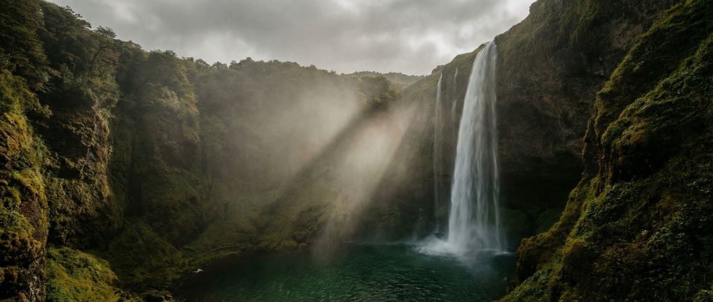 Waterfall plunging into a mossy gorge, mist rising into angled sunbeams, emerald pool at the base