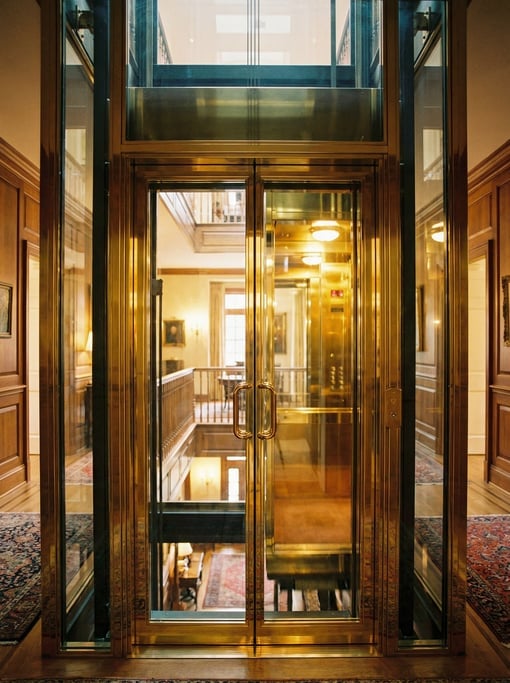 Tall narrow view of a home elevator with a glass door, polished brass interior