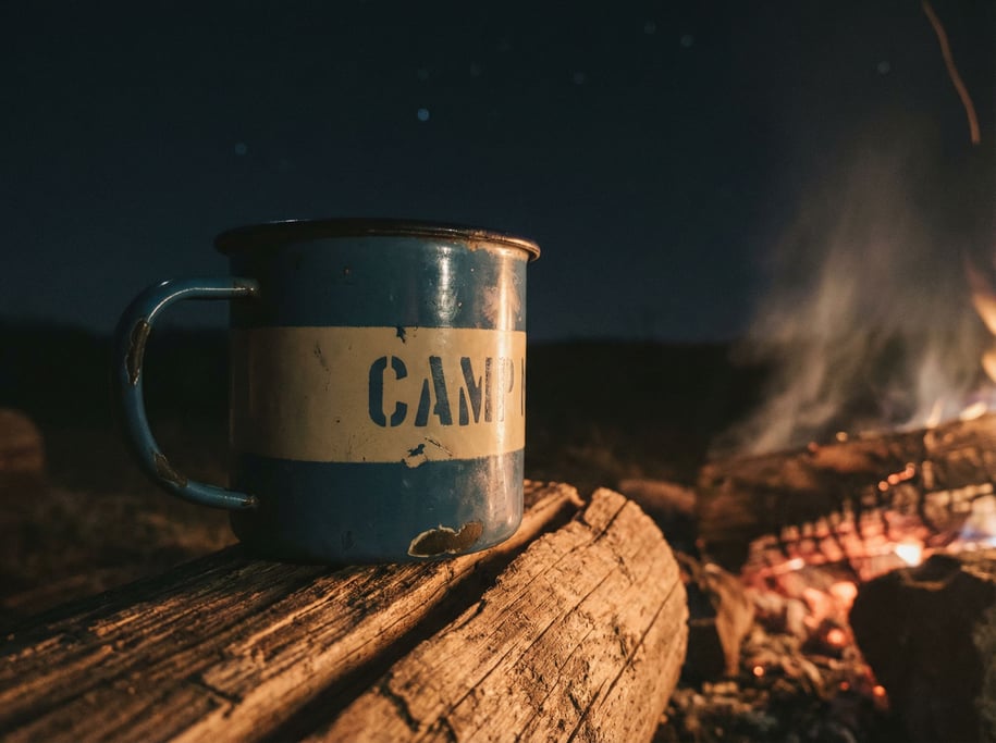 A vintage enamelware camping mug on a log next to a campfire, stars implied by the dark background