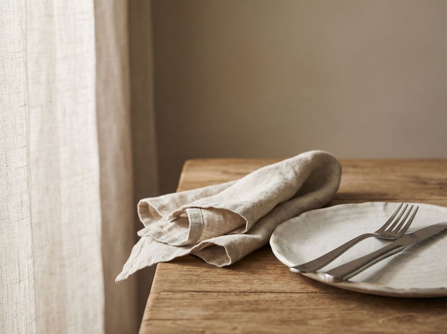 A linen napkin loosely folded beside a ceramic plate and steel cutlery, natural materials (e)
