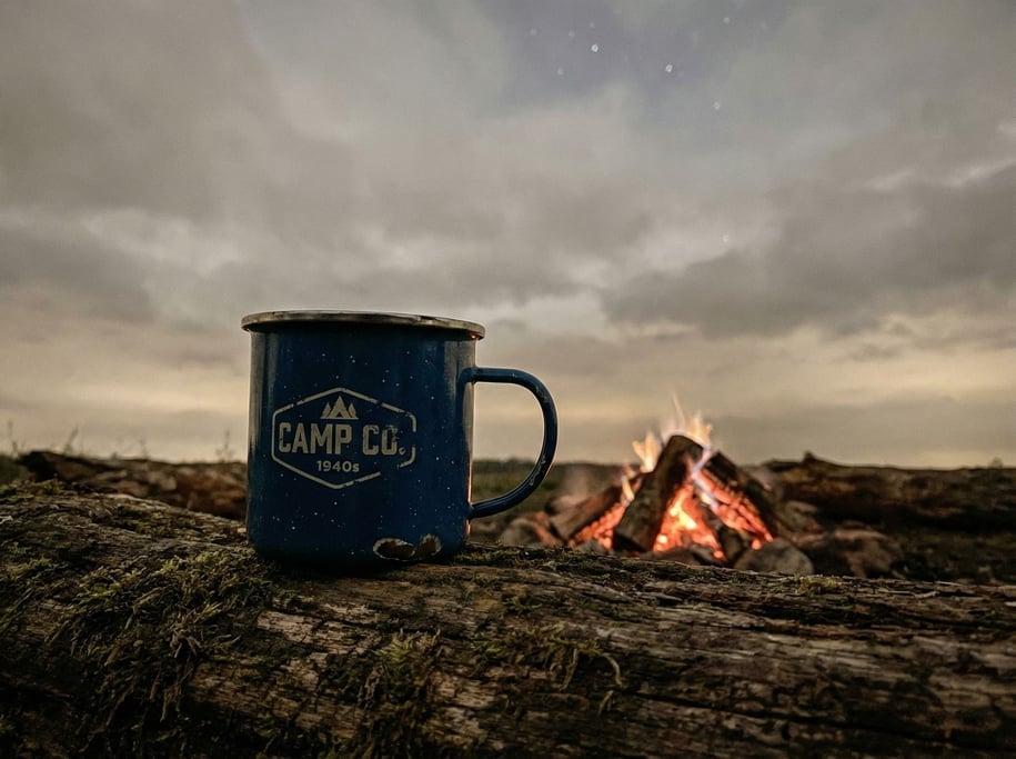 A vintage enamelware camping mug on a log next to a campfire, stars implied by the dark background (7esimwth)