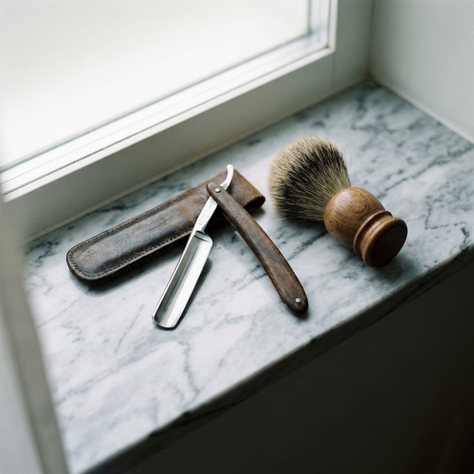 A vintage straight razor open on a marble shelf, chrome catching hard light, badger brush beside it (ocvybh9b)