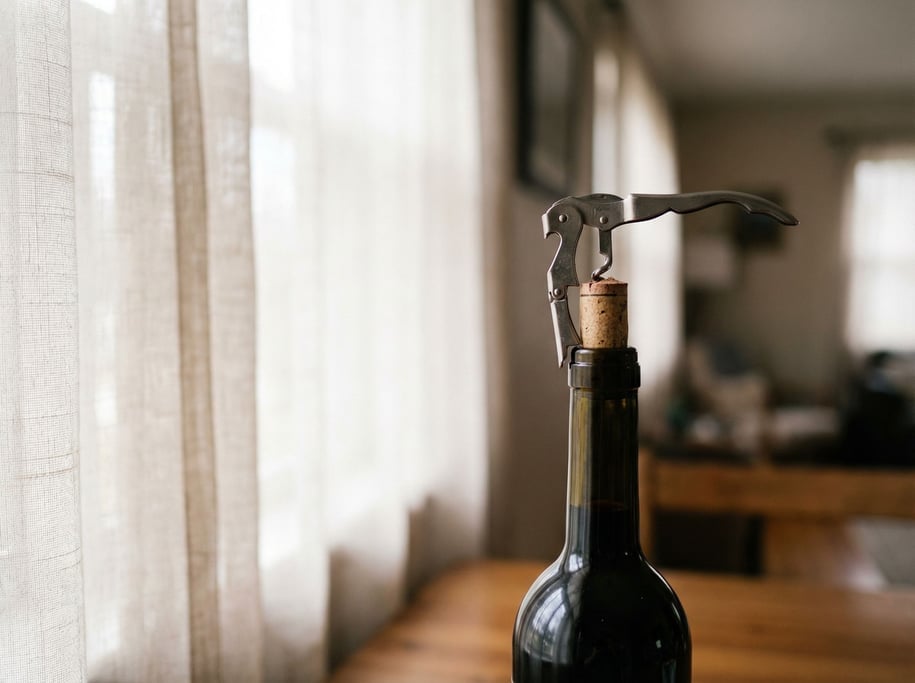 A corkscrew and wine cork mid-pull, red wine bottle in soft focus behind, ritual of opening (dhrkhex)