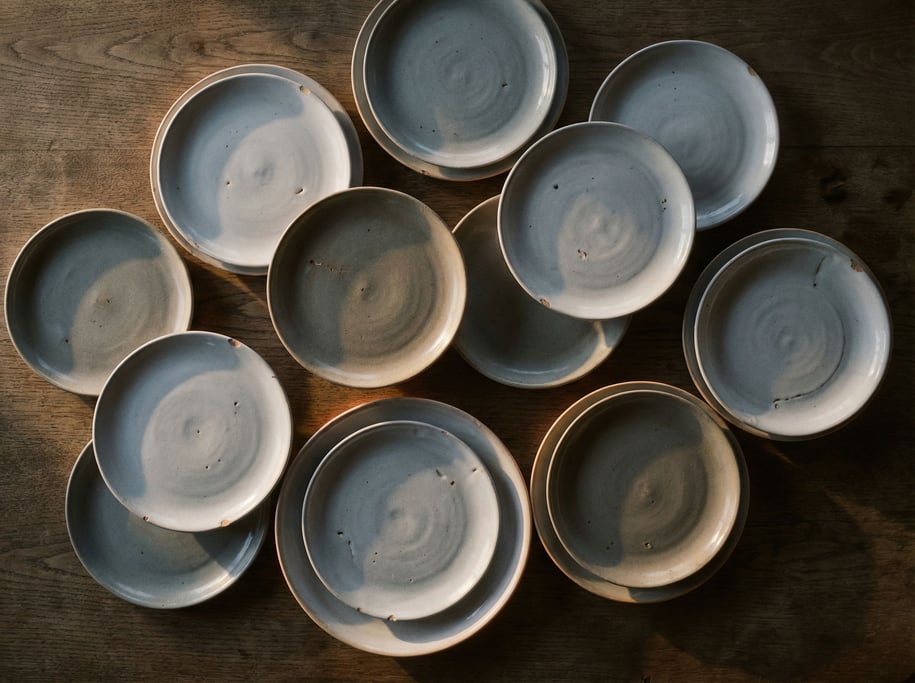 A stack of handmade ceramic plates in cool gray tones, slight imperfections visible, side lit (wtgirrw)