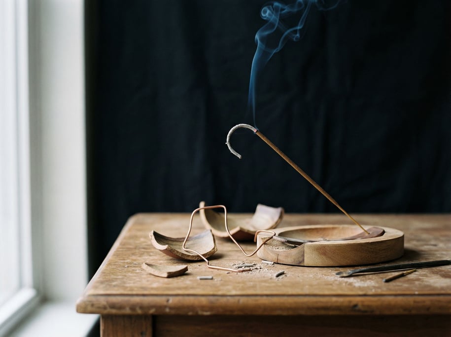 An incense holder with a smoking stick, ash curling in a thin line, single wisp rising against black (q3o3ufpi)