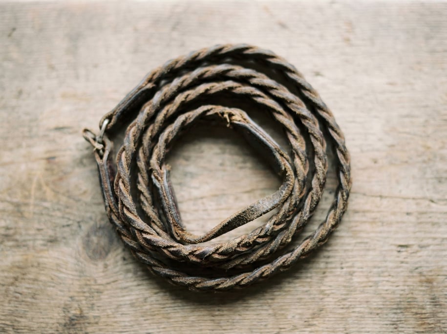 A vintage camera strap coiled on a wooden surface, braided leather showing years of neck wear (j39bgbak)