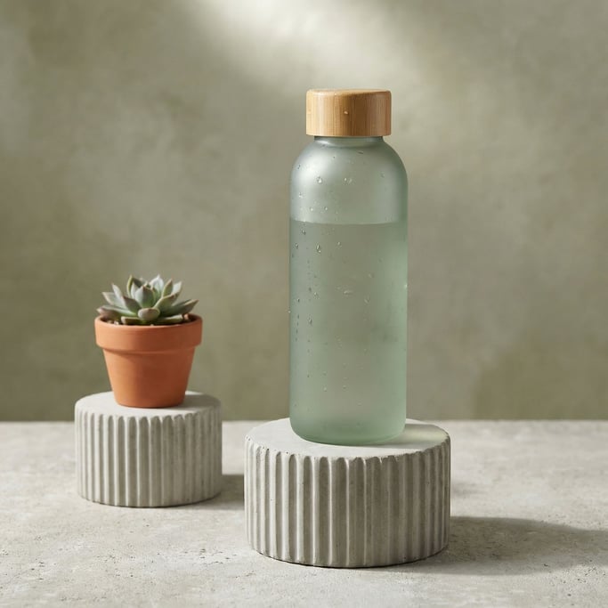 Sage green pervades: a frosted glass water bottle with a bamboo cap stands on a ribbed concrete cyli