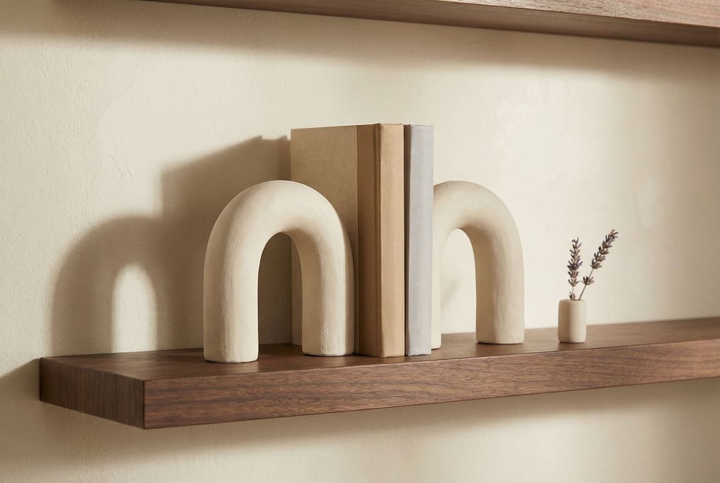 A pair of ceramic bookends in matte cream shaped as simple geometric arches on a dark walnut shelf section