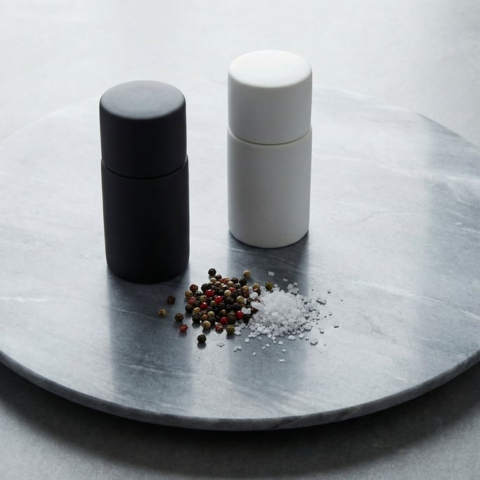 A pair of ceramic salt and pepper mills in contrasting matte white and matte black on a large piece of grey soapstone