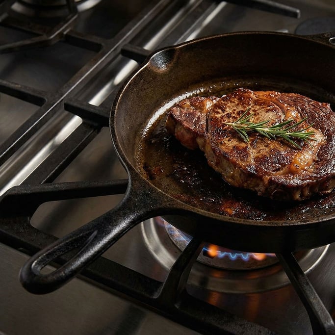 A cast iron skillet on a gas burner grate with a seared steak and rosemary sprig inside