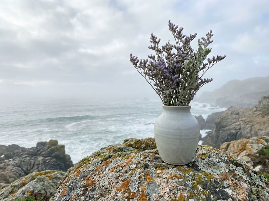 A ceramic vase in pale grey placed on a lichen-covered granite boulder by the coast
