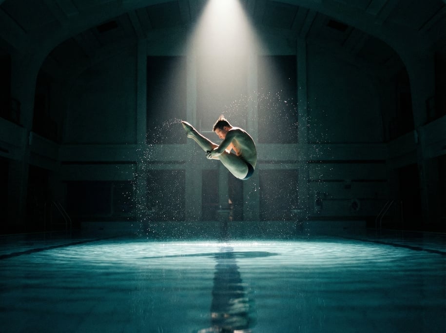 Cinematic shot of a diver in pike position mid-air above a calm pool surface