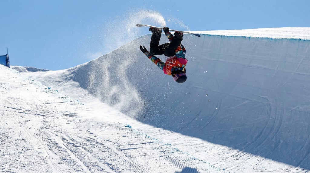 Snowboarder mid-grab trick in a halfpipe, fully inverted, snowy pipe walls and blue sky