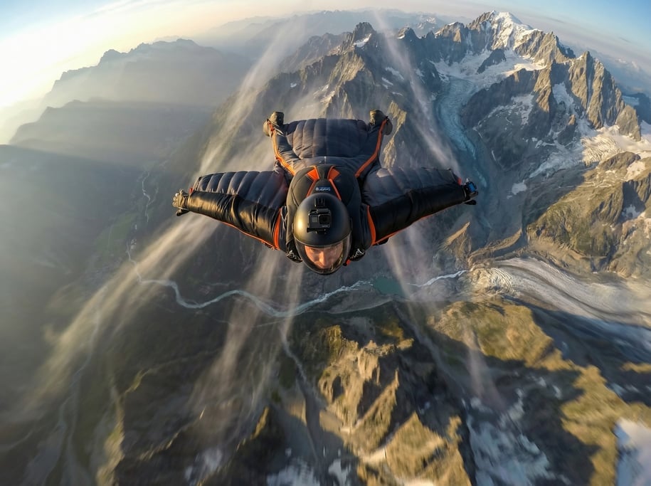 Wingsuit flyer in a spread-eagle position above a mountain landscape, wearing a dark suit