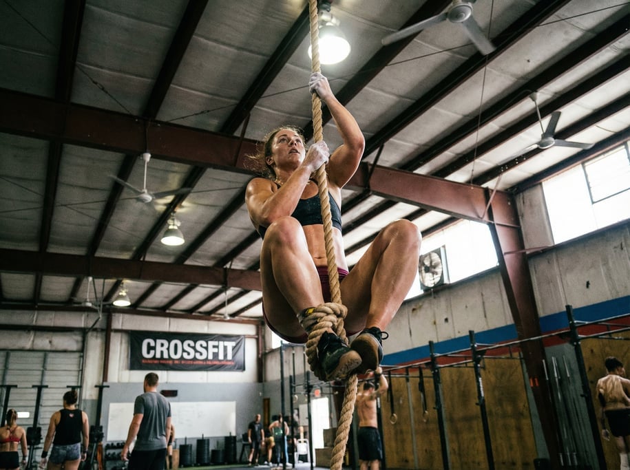 Athlete doing a rope climb in a CrossFit gym, halfway up a thick manila rope